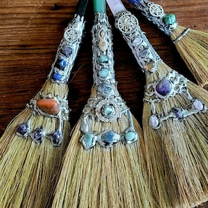 May include: Four small brooms with different colored crystal handles. Each handle is decorated with silver metalwork and charms. The brooms are made of natural fibers and have a rustic look.