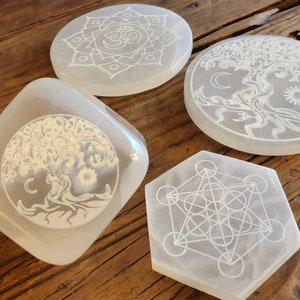 May include: Four white selenite charging plates with etched designs. The designs include a flower with an Om symbol in the center, a tree of life, a geometric pattern, and another tree of life.