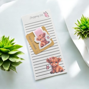May include: A white shopping list notepad with black horizontal lines and a pink butterfly illustration at the top. A square brown magnetic bookmark features a cartoon highland cow with a pink flower and a small butterfly on a pink background.
