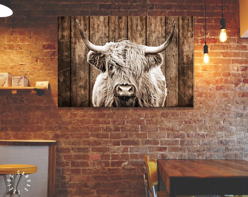 May include: A black and white photograph of a Highland cow with long, shaggy hair. The cow is looking directly at the camera with its head tilted slightly to the side. The background is a rustic wooden fence.
