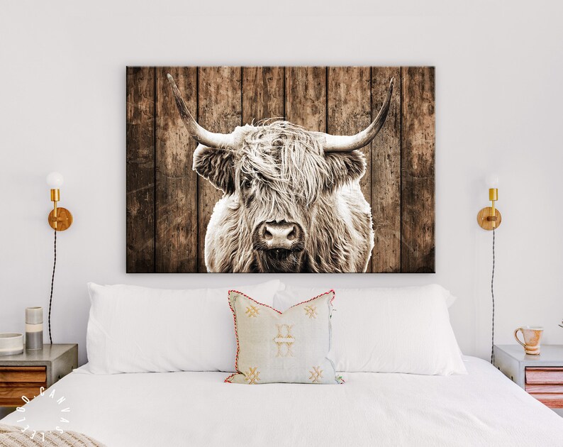 May include: A close-up of a Highland cow with long, shaggy fur and large horns. The cow is looking directly at the camera. The background is a rustic wooden wall.