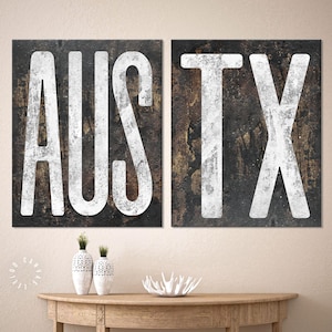 May include: Two black and white distressed wood signs with the words "AUST" and "TX" in white lettering. The signs are displayed on a wooden table with two white ceramic vases and a white decorative piece.