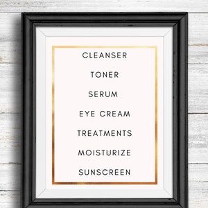 May include: A black framed print with a white background and gold lettering. The print lists the steps in a skincare routine: Cleanser, Toner, Serum, Eye Cream, Treatments, Moisturize, Sunscreen.
