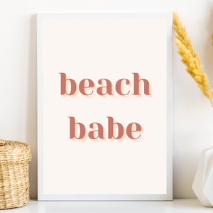 Puede incluir: Una impresi&oacute;n enmarcada en blanco con el texto "beach babe" en un color rosa melocot&oacute;n. La impresi&oacute;n est&aacute; sobre un fondo blanco y est&aacute; sobre una superficie blanca.
