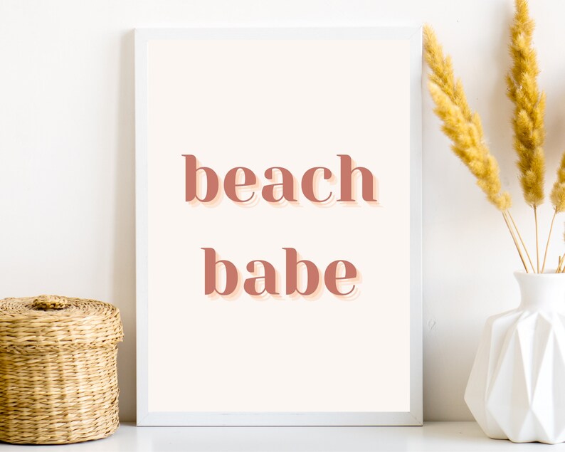Puede incluir: Una impresi&oacute;n enmarcada en blanco con el texto "beach babe" en un color rosa melocot&oacute;n. La impresi&oacute;n est&aacute; sobre un fondo blanco y est&aacute; sobre una superficie blanca.