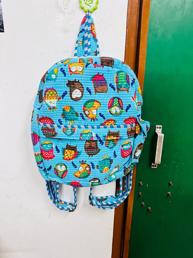 Puede incluir: Una mochila acolchada azul con estampado de b&uacute;hos coloridos. La mochila tiene un asa superior y correas ajustables.