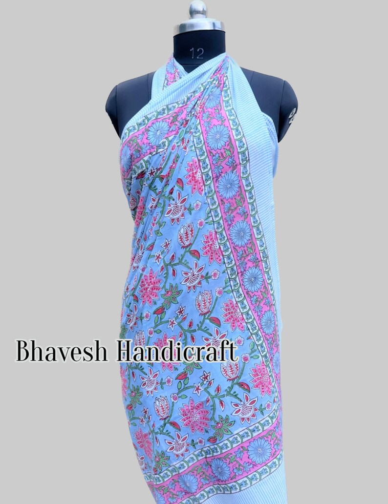 May include: A light blue cotton sarong with a floral print in shades of pink, green, and white. The sarong is tied around a mannequin and has a halter neck style. The text "Bhavesh Handicraft" is visible on the sarong.