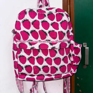 Puede incluir: Una mochila acolchada rosa y blanca con un estampado de fresas. La mochila tiene correas ajustables y un peque&ntilde;o bolsillo lateral.