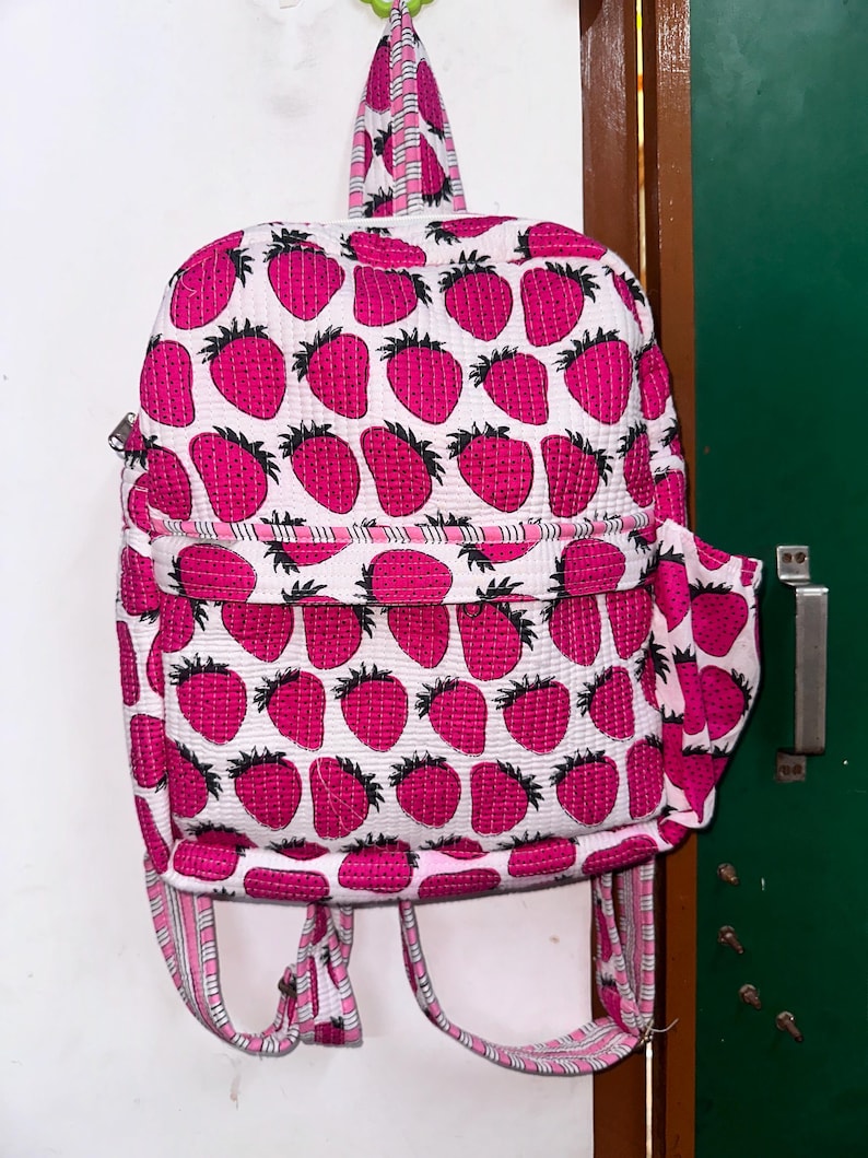 Puede incluir: Una mochila acolchada rosa y blanca con un estampado de fresas. La mochila tiene correas ajustables y un peque&ntilde;o bolsillo lateral.