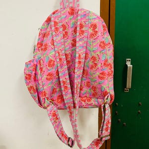 Puede incluir: Mochila rosa floral con rayas blancas en las correas. La mochila tiene un bolsillo grande en la parte delantera y un bolsillo m&aacute;s peque&ntilde;o en el lateral.