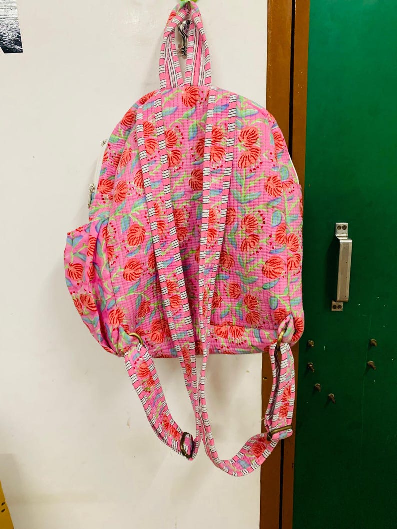 Puede incluir: Mochila rosa floral con rayas blancas en las correas. La mochila tiene un bolsillo grande en la parte delantera y un bolsillo m&aacute;s peque&ntilde;o en el lateral.