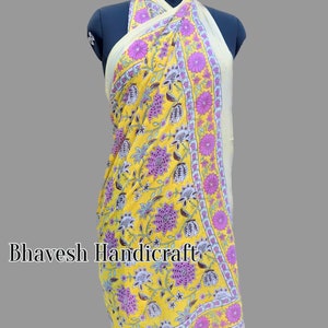 May include: A yellow sarong with a floral print in shades of pink and white. The sarong has a border with a repeating pattern of pink flowers and a white and yellow geometric design. The text "Bhavesh Handicraft" is visible in the lower left corner of the image.