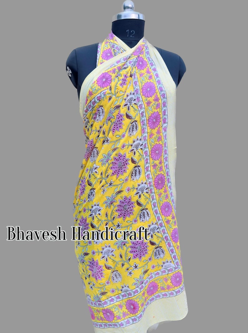 May include: A yellow sarong with a floral print in shades of pink and white. The sarong has a border with a repeating pattern of pink flowers and a white and yellow geometric design. The text "Bhavesh Handicraft" is visible in the lower left corner of the image.