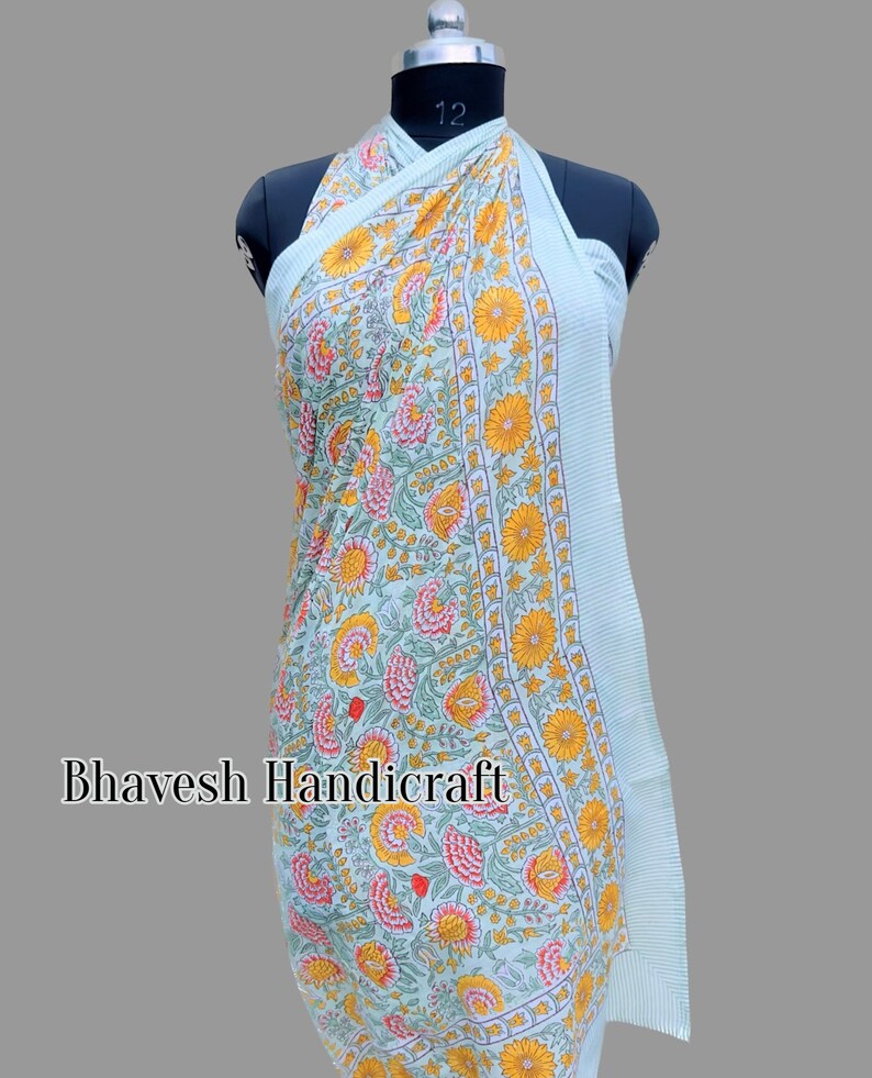 May include: A light blue sarong with a floral print featuring yellow, pink, and green flowers. The sarong is draped over a mannequin and has a white stripe border.