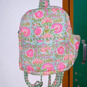 Puede incluir: Una mochila azul claro con estampado floral de flores rosas y verdes. La mochila tiene correas ajustables y cierre de cremallera.
