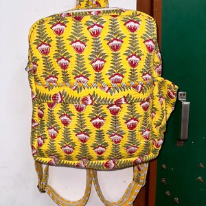 Puede incluir: Una mochila acolchada amarilla con un estampado floral en rojo y verde. La mochila tiene correas ajustables y un cierre de cremallera.