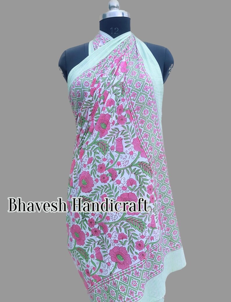 May include: A white cotton sarong with a pink floral print and green geometric border. The sarong is draped over a mannequin and is styled as a dress. The text "Bhavesh Handicraft" is visible on the sarong.