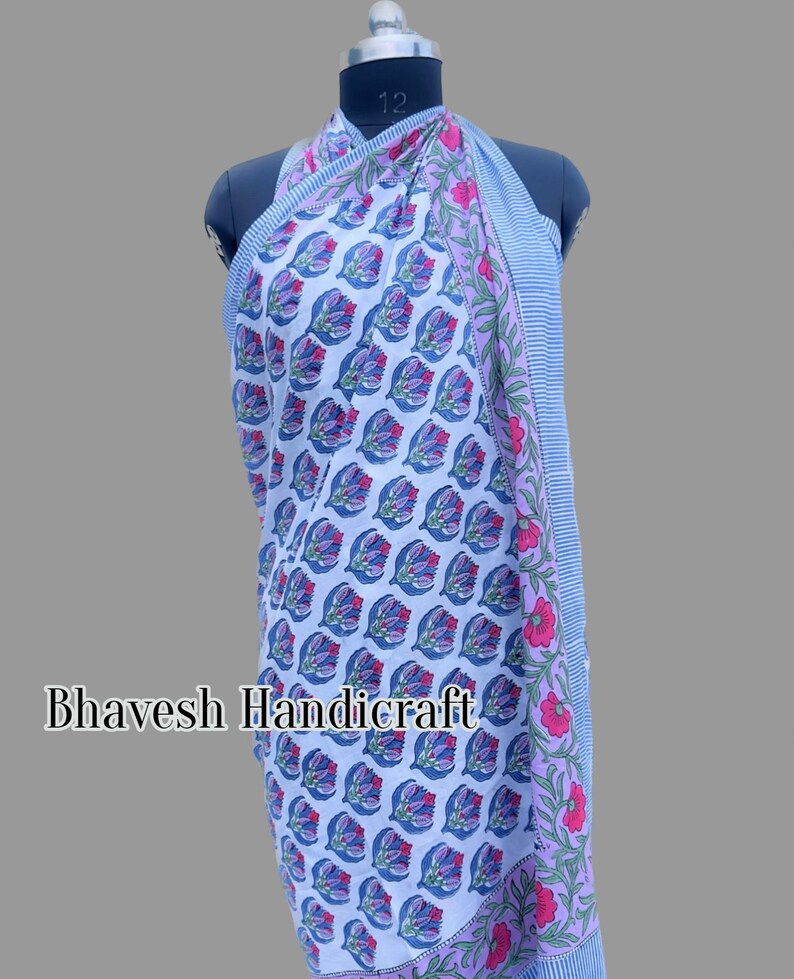 May include: A blue and white cotton sarong with a floral print. The sarong has a pink and white border with a striped pattern. The sarong is draped over a mannequin.