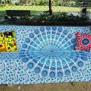Puede incluir: Un tapiz mandala blanco y azul con un diseño de pavo real. El tapiz está drapeado sobre un sofá con dos cojines coloridos.