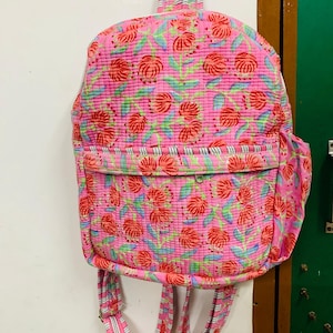 Peut inclure: Un sac à dos rose avec un motif floral composé de fleurs rouges et de feuilles vertes. Le sac à dos a des bretelles réglables avec des rayures blanches et noires.