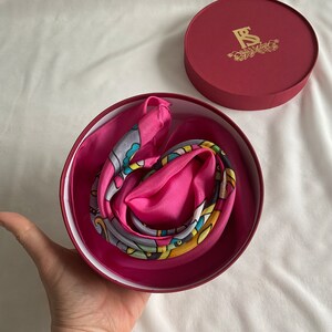 May include: A pink and multi-colored patterned silk scarf is folded and placed inside a red circular box with a gold logo that says "FS".