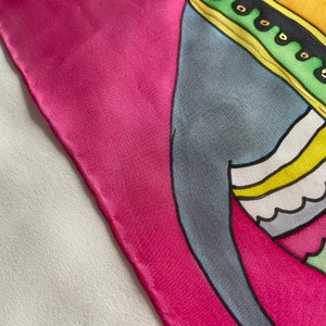 May include: A pink silk scarf with a colorful abstract design. The design features a blue, yellow, and green figure with a gold trim.