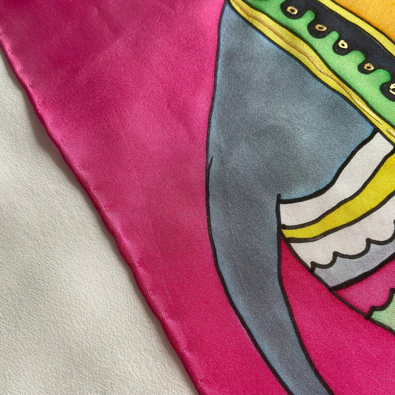May include: A pink silk scarf with a colorful abstract design. The design features a blue, yellow, and green figure with a gold trim.