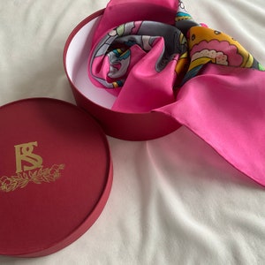 May include: A red round box with a gold logo of the letters "RS" surrounded by a wreath. The box is open and a pink silk scarf with a colorful abstract design is inside.
