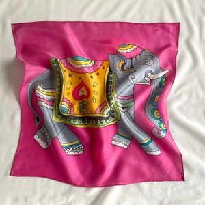 May include: A pink silk scarf with a colorful illustration of an elephant wearing a decorative headdress. The elephant is decorated with intricate patterns and bright colors.