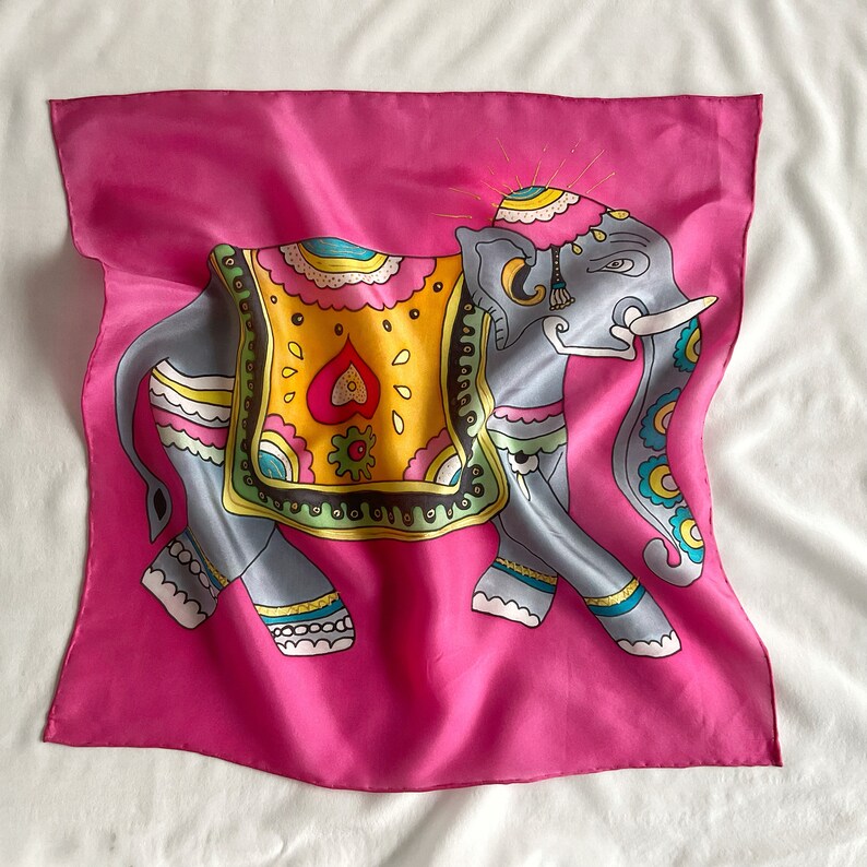 May include: A pink silk scarf with a colorful illustration of an elephant wearing a decorative headdress. The elephant is decorated with intricate patterns and bright colors.
