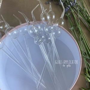 May include: Clear glass applicators with bulb-shaped reservoirs and curved tips are arranged in a light pink bowl. The text "GLASS APPLICATOR for gutta" is visible. Lavender sprigs are in the background.