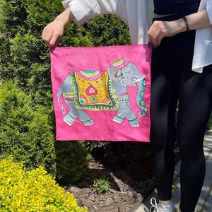 May include: A pink silk scarf with a colorful illustration of an elephant. The elephant is decorated with intricate patterns and has a blue and green floral design on its head.