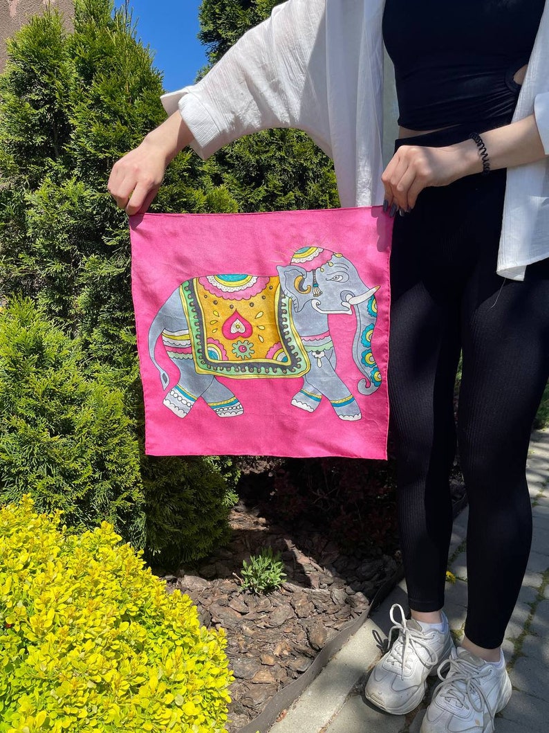May include: A pink silk scarf with a colorful illustration of an elephant. The elephant is decorated with intricate patterns and has a blue and green floral design on its head.