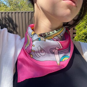 May include: A pink silk scarf with a colorful elephant design. The scarf is tied around the neck of a person.