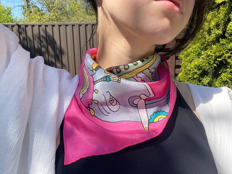 May include: A pink silk scarf with a colorful elephant design. The scarf is tied around the neck of a person.