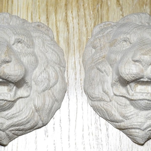 Può includere: Due sculture in legno a forma di testa di leone. I leoni sono scolpiti in legno chiaro, con criniere dettagliate, bocche aperte e denti visibili. Le sculture sono esposte su uno sfondo chiaro con venature del legno, che mette in risalto la naturale consistenza del legno.