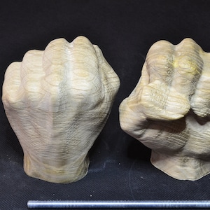 May include: Two wooden sculptures of clenched fists, side by side, with visible wood grain. The fists are light brown with hints of yellow and sit on a black surface. A metal rod is in the foreground.