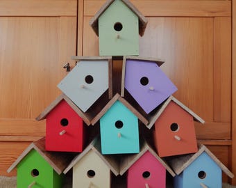 Könnte beinhalten: Ein pyramidenförmiger Stapel aus zehn Holzvogelhäusern in verschiedenen Farben, darunter Hellgrün, Grau, Lila, Rot, Türkis, Orange, Limettengrün, Creme, Pink und Hellblau. Jedes Vogelhaus hat ein rundes Einflugloch und eine kleine Sitzstange.