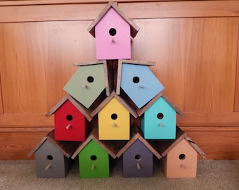 Peut inclure: Une pyramide de nichoirs en bois peints de couleurs vives. Chaque nichoir est peint d'une couleur différente : rose, vert, bleu, rouge, jaune, gris et pêche. Ils ont un trou d'entrée rond et un perchoir en bois.