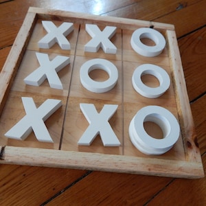 May include: A wooden tic-tac-toe game with a light brown frame and a grid of nine squares. The game pieces are white, with five X's and four O's. The game is set on a wooden surface.