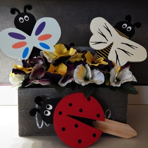 May include: A gray wooden box with a floral arrangement of white, purple, and yellow flowers.  The box has three wooden decorations: a butterfly with orange, blue, and purple accents, a bee with black and yellow stripes, and a ladybug with red and black spots.