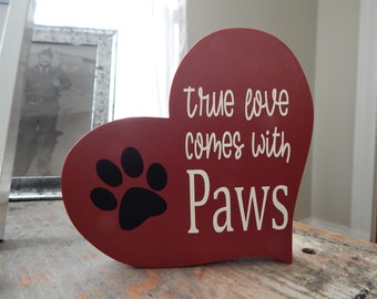Può includere: Un cartello di legno rosso a forma di cuore con un'impronta di zampa nera e il testo "true love comes with paws" in bianco.