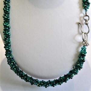 May include: Emerald green beaded necklace displayed on a white mannequin bust. The necklace is made of small, iridescent beads, and features a silver-toned clasp. The beads are tightly strung together, creating a textured, rope-like appearance.