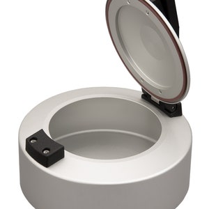 Puede incluir: Un recipiente redondo de plata con una tapa con bisagras. La tapa está abierta y tiene un asa negra. El recipiente tiene una junta de goma negra alrededor de la tapa.