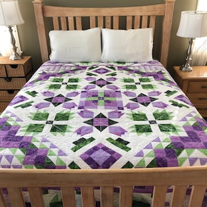 May include: A white quilt with a purple and green geometric pattern. The quilt is on a wooden bed frame with a white headboard.