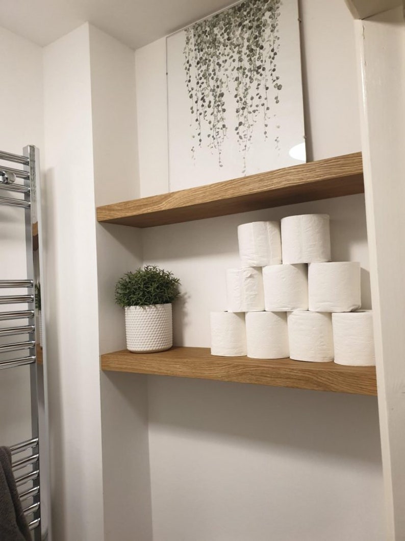 May include: A bathroom scene featuring two wooden shelves. The top shelf holds a framed artwork with a green plant design. The bottom shelf displays a stack of white toilet paper rolls and a small potted plant in a white textured pot.