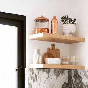 May include: Two wooden shelves display kitchen decor. The top shelf holds a copper-toned jar, bottle, and a white pot with greenery. The bottom shelf features a white vase, wooden cutting boards, white bowls, and glasses.
