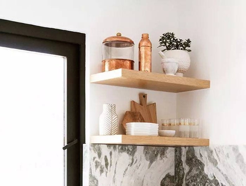 May include: Two wooden shelves display kitchen decor. The top shelf holds a copper-toned jar, bottle, and a white pot with greenery. The bottom shelf features a white vase, wooden cutting boards, white bowls, and glasses.