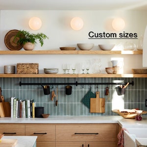 May include: A modern kitchen with light wood cabinets, open shelving, and a teal tile backsplash. The shelves display bowls, plants, and kitchenware. A black metal rod holds utensils and cutting boards. The text "Custom sizes" is visible.