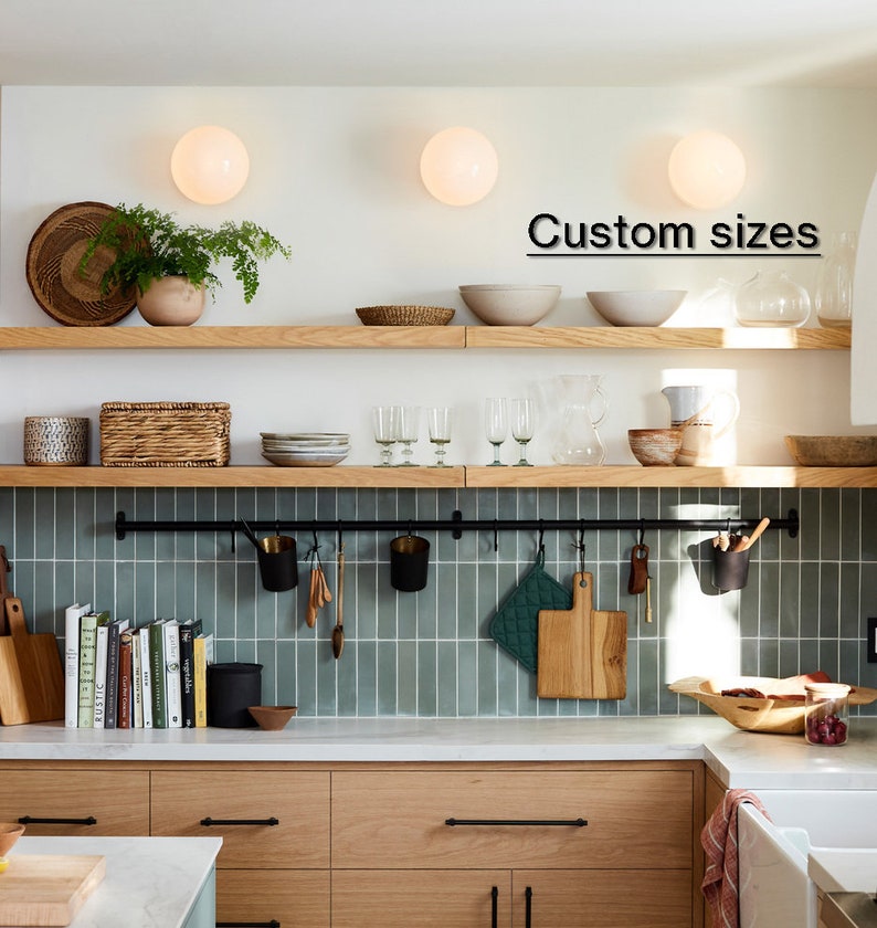 May include: A modern kitchen with light wood cabinets, open shelving, and a teal tile backsplash. The shelves display bowls, plants, and kitchenware. A black metal rod holds utensils and cutting boards. The text "Custom sizes" is visible.